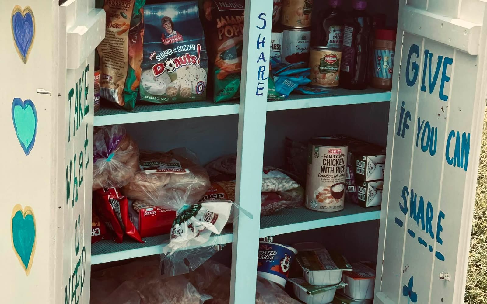 Inside a white Blessing Box painted with hearts, its shelves stocked with bread, canned goods and snacks